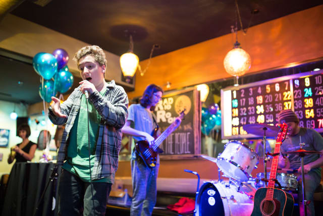 Young male singer performing on stage with microphone, accompanied by a bassist and drummer, in a lively venue decorated with balloons, capturing the essence of the Love Bomb event supporting My Voice Music.